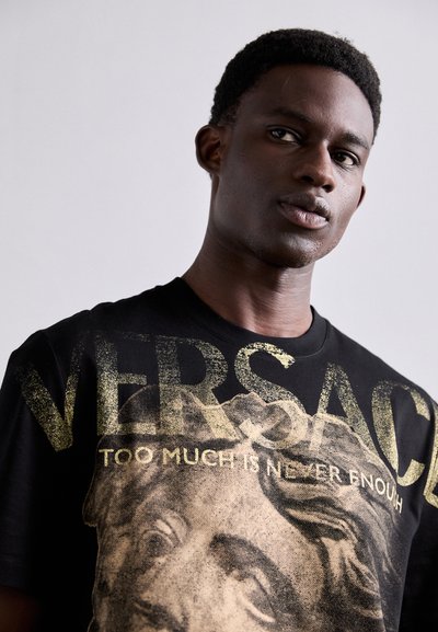 T-shirt noir avec impression graphique dorée d'un visage texturé et texte "VERSACE". Design à col rond avec manches courtes.