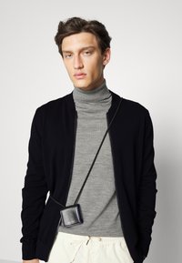 Black zip-up bomber jacket over a gray turtleneck, with a small, shiny black crossbody pouch on a black strap.