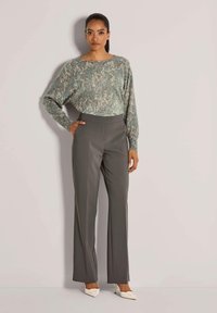 Vrouw staat tegen de muur met een grijze, patroonrijke lange mouwtop, hoge taille grijze wijde broek en witte puntige hakken.