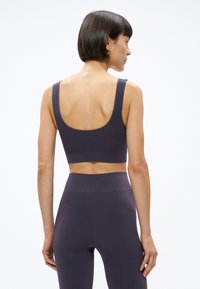 Žena s krátkymi tmavými vlasmi, oblečená v zodpovedajúcej tmavofialovej športovej podprsenke a legínach s vysokým pásom, zobrazená zozadu na bielom pozadí.