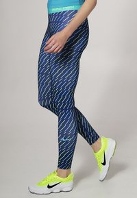 Leggings a fantasia blu e verde con vita alta, realizzati in materiale elastico. Abbinati a scarpe da ginnastica gialle vivaci.