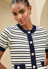 Cardigan lavorato a maglia con righe orizzontali blu e crema, maniche corte, collo rotondo e chiusura con bottoni dorati, completo di tasche.