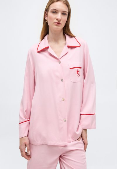 Γυναίκα που φοράει ροζ πυτζάμες με κουμπιά, λεπτομέρειες σε κόκκινο και κέντημα αστακού στην τσέπη, στέκεται μπροστά σε απλό λευκό φόντο.