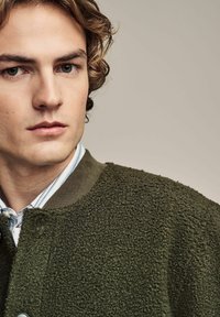 Men's green textured jacket with a ribbed collar, featuring a button closure. Worn over a patterned blue and white shirt.