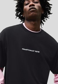 Tricou negru cu text alb brodat "CHAOTICALLY CUTE". Purtați peste o cămașă cu mâneci lungi roz deschis. Textură netedă, croială lejeră.