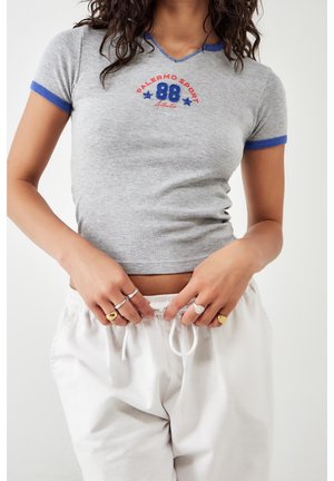 Kobieta nosząca dopasowany szary krótki t-shirt z niebieską lamówką i napisem „Palermo Sport 88”, regulująca sznurek w białych spodniach.
