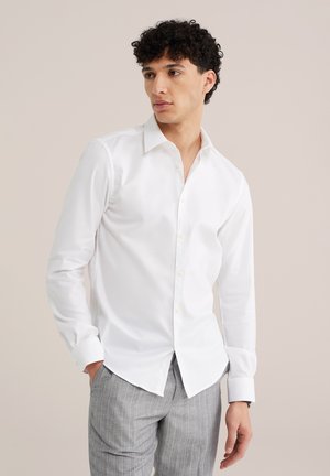 Mand med krøllet hår iført en hvid skjorte med knapper og grå bukser med striber, stående med den ene hånd i lommen.