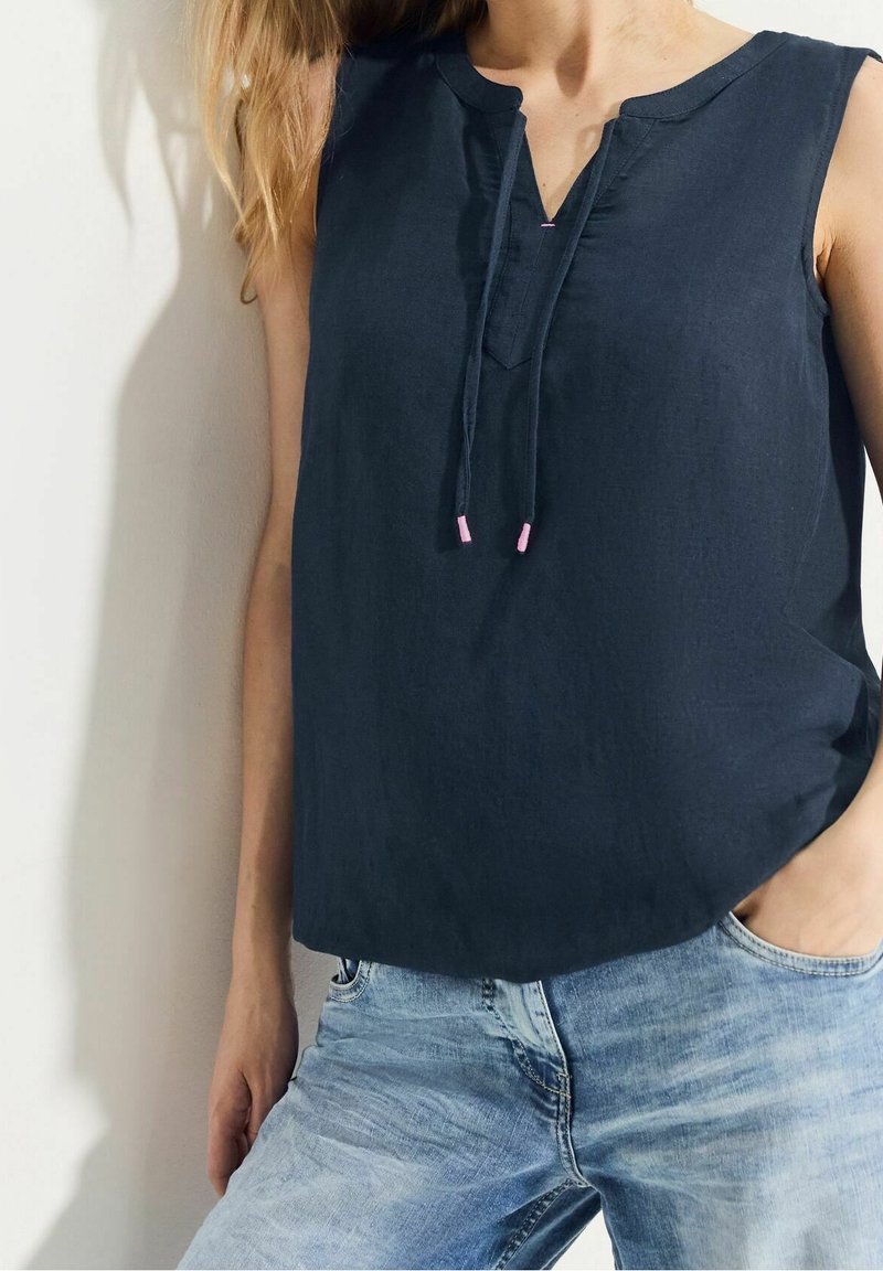 Femme portant un haut sans manches bleu marine avec un détail noué à l'avant, associé à un jean délavé bleu clair, debout devant un mur blanc.