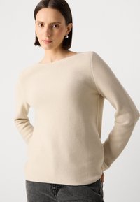 Suéter de malha bege com decote barco e mangas longas, apresentando uma textura suave e um ajuste descontraído. Usado com jeans cinza escuro.