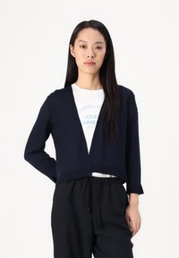 Cardigan cropped navy con maniche lunghe, texture a maglia, design aperto sul davanti, indossato sopra una t-shirt grafica bianca e pantaloni neri con cordino.