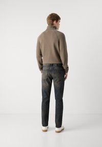 Suéter de punto marrón con cuello alto, combinado con jeans azules desgastados y zapatillas blancas con suela de goma. Fondo liso.