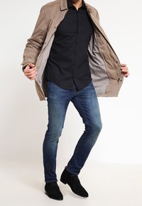Light brown jacket with a grey interior, black button-up shirt, dark blue jeans, and black ankle boots. Standing pose, jacket open.