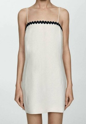 Femme portant une mini-robe blanche sans manches avec de fines bretelles et un bord ondulé noir le long de l'encolure sur un fond uni.