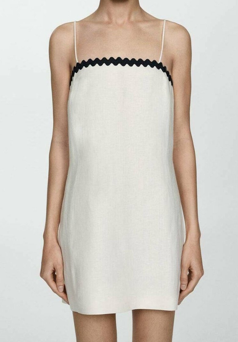 Femme portant une mini-robe blanche sans manches avec de fines bretelles et un bord ondulé noir le long de l'encolure sur un fond uni.
