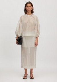 Leichtes, transparentes beiges Oberteil mit Puffärmeln und rundem Ausschnitt, kombiniert mit einem passenden Midikleid. Hält eine schwarze Clutch.