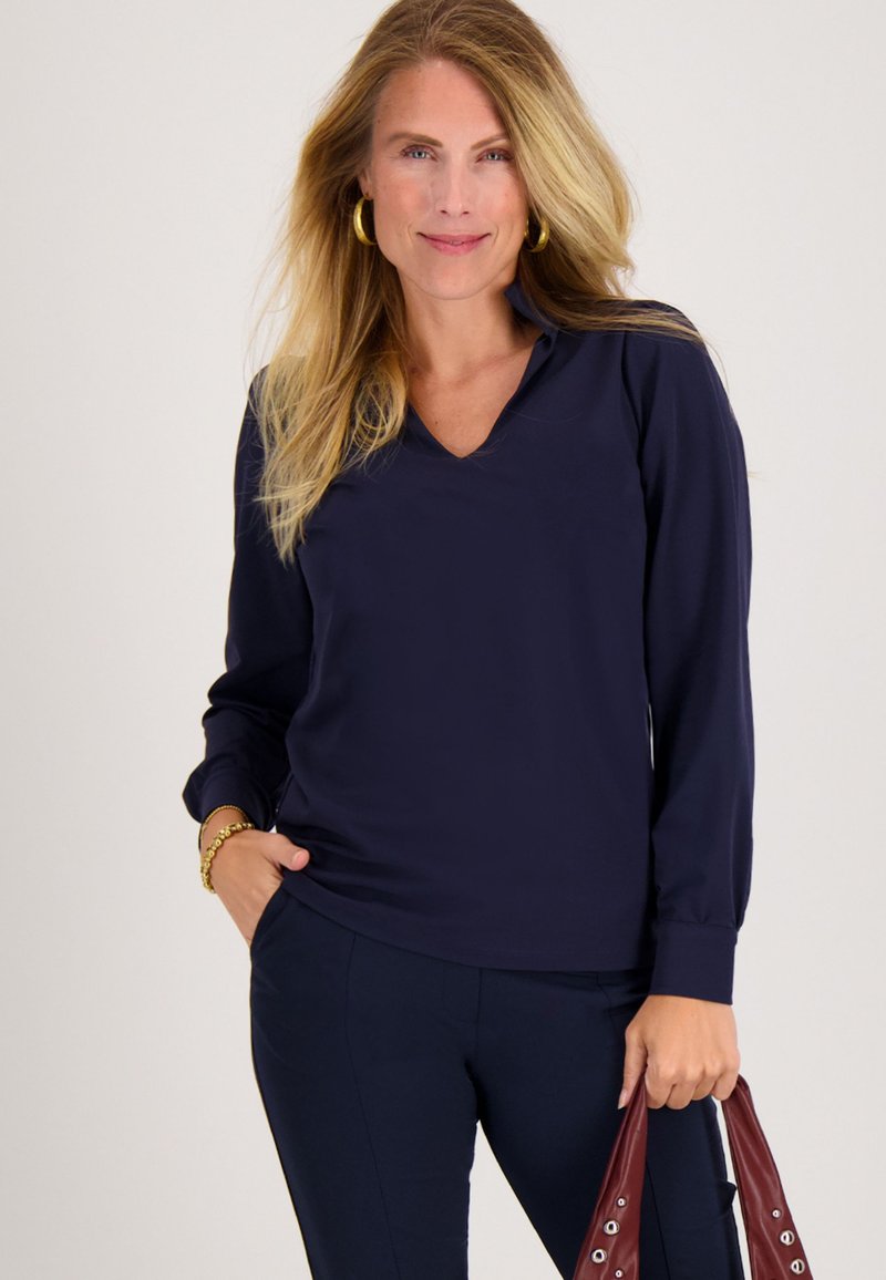 Vrouw met lang blond haar, gekleed in een marineblauw shirt met lange mouwen en een broek, houdt een kastanjebruine handtas vast en glimlacht tegen een effen achtergrond.