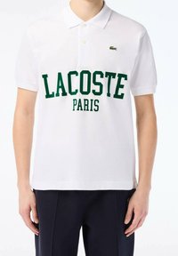 Person wearing white Lacoste polo shirt with green "LACOSTE PARIS" text and small crocodile logo on chest, paired with dark pants.