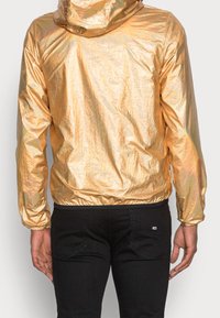 Zlatá reflexní lehká bunda s kapucí, elastickými manžetami a lemem. Nositelná přes černé kalhoty. Mačkaná textura zvyšuje vizuální hloubku.
