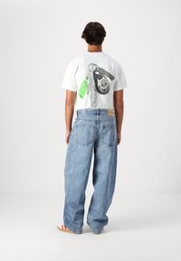 T-shirt blanc avec un graphisme d'outils dans le dos, associé à un jean en denim bleu clair surdimensionné et des chaussures beiges avec des rayures rouges.