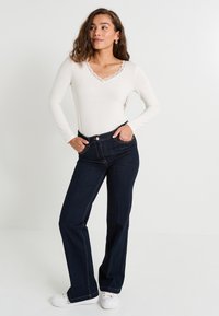 Femme aux cheveux ondulés portant un haut blanc avec des bordures en dentelle, un jean bleu foncé et des baskets blanches, se tenant avec les mains dans les poches devant un fond uni.