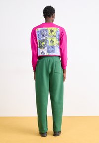 Langsleeve roze top met een geblokte achterkant in blauw, groen en geel. Gecombineerd met groene joggingbroek en zwarte schoenen.