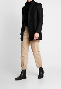 Manteau en laine noire avec un col montant, une fermeture boutonnée et deux poches latérales. Associé à un pantalon chino beige fuselé et des bottines noires.