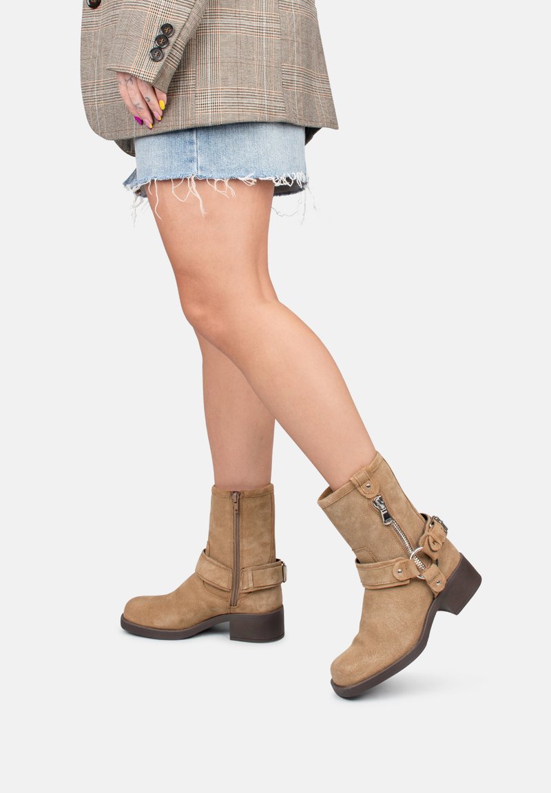 Botines de ante en color marrón con hebillas y cremalleras. Tacón grueso de color marrón. Combinados con un blazer de cuadros y shorts de denim, mostrando un estilo en capas.