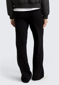 Persona che indossa pantaloni larghi neri, sneakers bianche, una camicia bianca e un giubbotto trapuntato nero, in piedi con le mani rilassate ai lati.