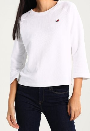 Kobieta ubrana w białą koszulę z długim rękawem z małym logo Tommy Hilfiger na piersi i ciemnoniebieskie dżinsy.