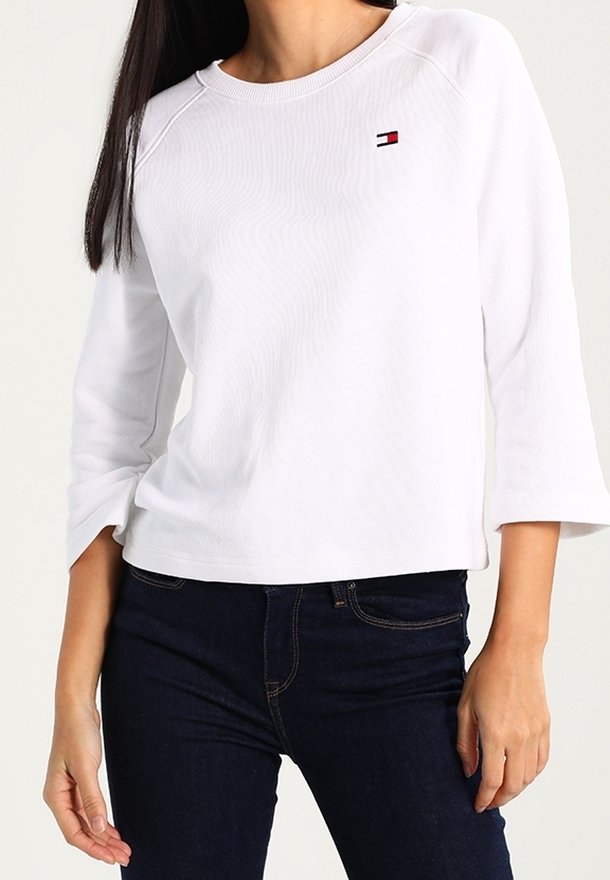 Femme portant une chemise blanche à manches longues avec un petit logo Tommy Hilfiger sur la poitrine et un jean bleu foncé.