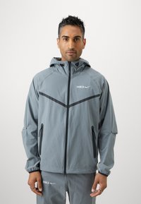 Γκρίζα, ελαφριά τζάκετ με κουκούλα, πλήρη φερμουάρ μπροστά, μαύρες λεπτομέρειες, δύο πλευρικές τσέπες και λογότυπα της BARCA και της Nike στο στήθος.