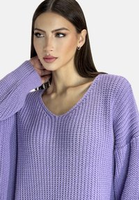 Lavendelfarbenes Strickpullover mit V-Ausschnitt, breiten Ärmeln und einem strukturierten Muster. Der Stoff wirkt weich und leicht.