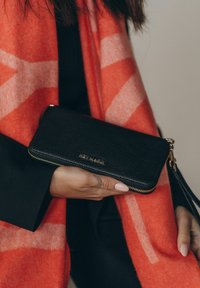 Zwarte leren portemonnee met een gestructureerd oppervlak, gouden rits en reliëfbranding. Vastgehouden met een felrode sjaal met een geometrisch patroon.
