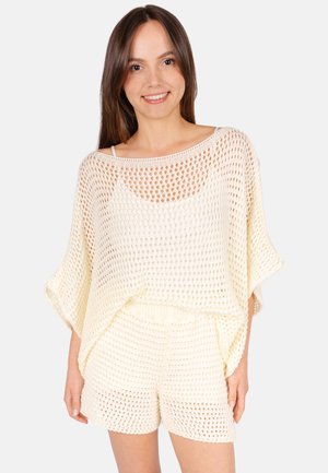 Femme aux longs cheveux bruns portant un haut blanc en maille ample et un short assorti, souriante devant un fond clair uni.