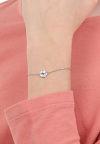 Delicado brazalete de plata en la muñeca con cuatro charms en forma de corazón, uno de ellos engastado con pequeños cristales, llevado sobre una camiseta de manga larga rosa.