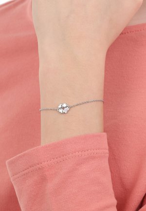 Delicado brazalete de plata en la muñeca con cuatro charms en forma de corazón, uno de ellos engastado con pequeños cristales, llevado sobre una camiseta de manga larga rosa.