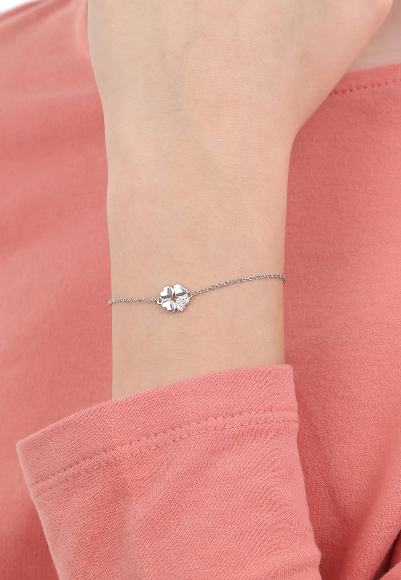 Delicado brazalete de plata en la muñeca con cuatro charms en forma de corazón, uno de ellos engastado con pequeños cristales, llevado sobre una camiseta de manga larga rosa.