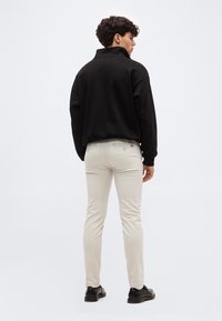 Νέος άνδρας που στέκεται και κοιτάζει μακριά, φορώντας μαύρο μακρυμάνικο μπλουζάκι, μπεζ στενά παντελόνια και μαύρα παπούτσια σε απλό ανοιχτό φόντο.