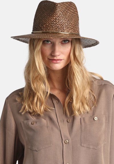 Blonde vrouw met los haar die een bruine geweven hoed met brede rand en een taupekleurig overhemd met borstzakken draagt.