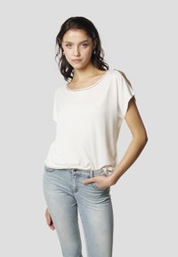 Valge lühikeste varrukatega pluus, millel on kaunistatud helmekestega ümara kaelusega, kombineeritud helesiniste kitsaste teksadega, esile tõstetud neutraalsel taustal.