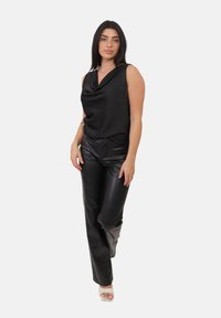 Donna in piedi con un top drappeggiato senza maniche nero, pantaloni in pelle neri e sandali con tacco aperti bianchi, davanti a uno sfondo neutro.