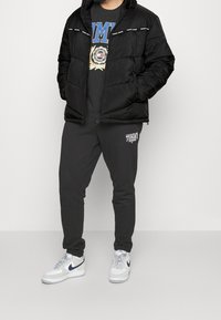 Černá péřová bunda s kapucí, tričko s kulatým výstřihem a grafikou, šedé tepláky s logem, bílé tenisky s námořnickými doplňky, pohodlný střih.