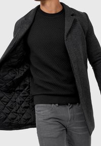 Grauer Wollmantel mit strukturiertem schwarzem Innenfutter, getragen über einem schwarzen Stepppullover. Der Mantel hat einen Reverskragen und eine schlanke Silhouette.