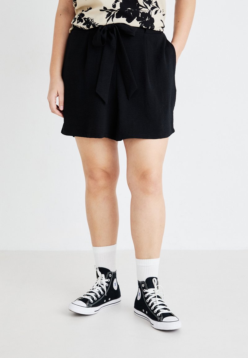 Pantalones cortos negros con textura y un lazo en la cintura, combinados con unas zapatillas altas negras y calcetines blancos. Presente en la imagen una blusa floral.