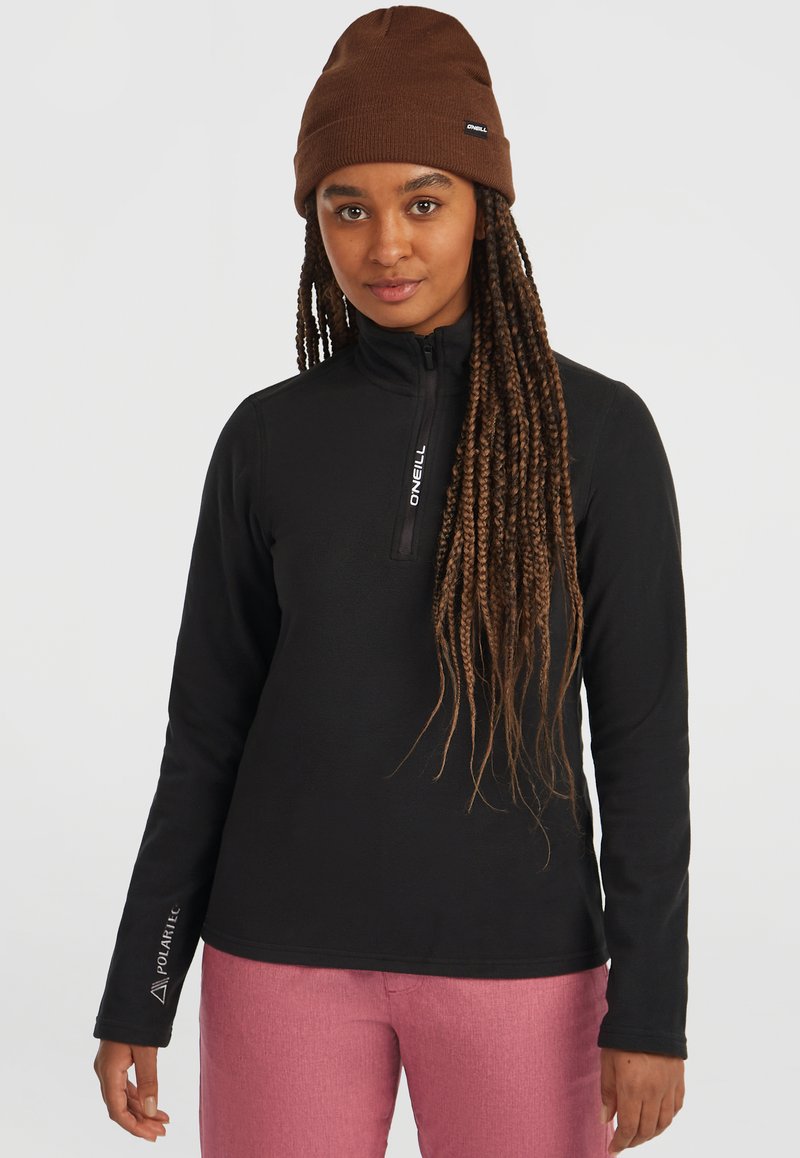 Zwarte fleecejack met een half-rits aan de voorkant, met een fleece textuur en een wit logo op de linkerarm. Gecombineerd met roze broek.