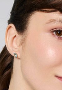 Złote kolczyki z prostokątnym niebieskim kamieniem, ozdobione mniejszymi, przezroczystymi kamieniami. Projekt jest elegancki, w kontekście jasnej karnacji.