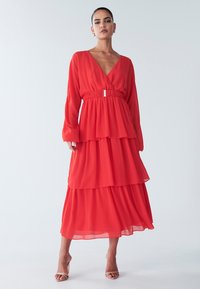 Abito rosso a strati realizzato in tessuto leggero, con maniche lunghe, scollo a V profondo e vita con cintura. Texture liscia e design svolazzante.