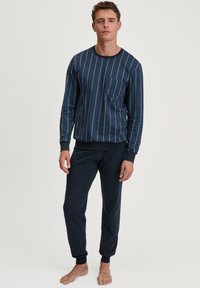 Langarmige marineblauwe top met verticale patronen in lichterblauw en oranje, voorzien van een zak; gecombineerd met donker marineblauwe broeken met nauwsluitende boorden.