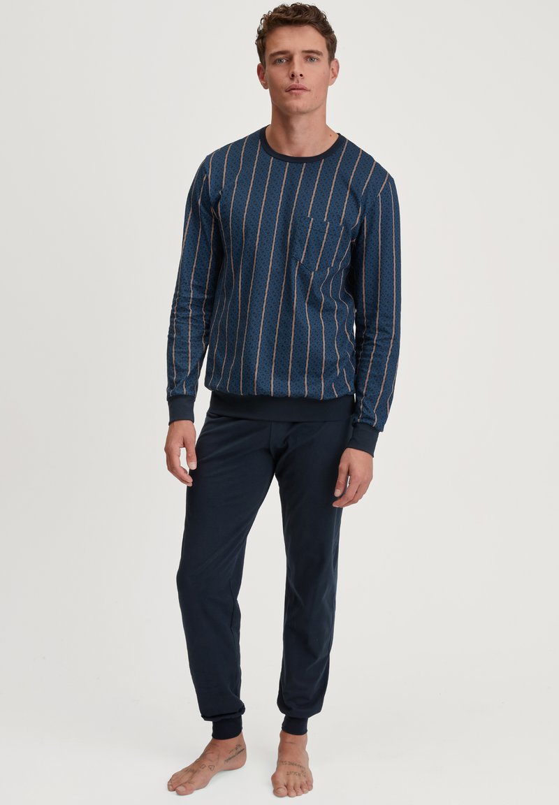 Langarmige marineblauwe top met verticale patronen in lichterblauw en oranje, voorzien van een zak; gecombineerd met donker marineblauwe broeken met nauwsluitende boorden.