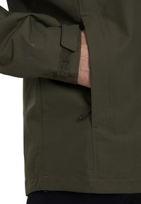 Närbild av en persons hand som vilar i fickan på en vattentät, olivgrön jacka med justering för kardborreförslutning vid ärmen.